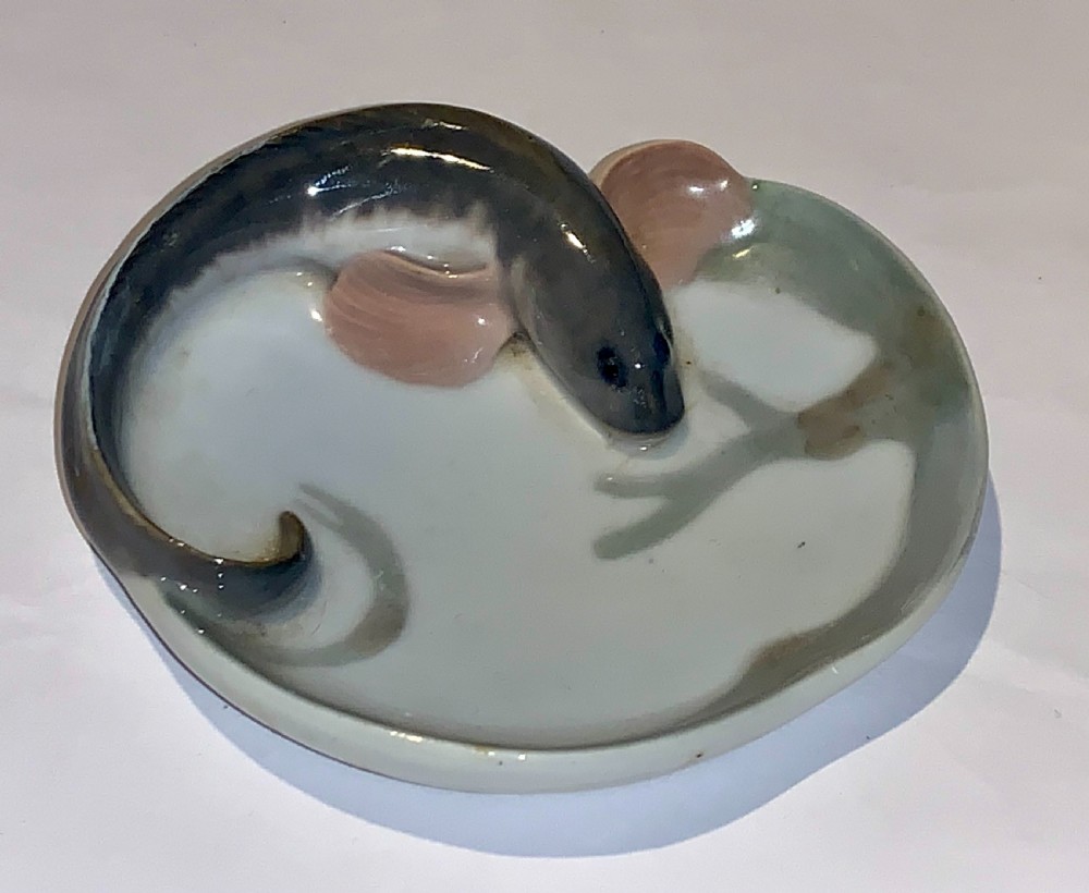 A small circular porcelain dish featuring a sculpted grey fish with pink fins moulded along the curved rim against an off-white background. View II