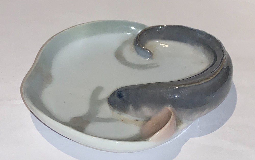 A small circular porcelain dish featuring a sculpted grey fish with pink fins moulded along the curved rim against an off-white background. View III