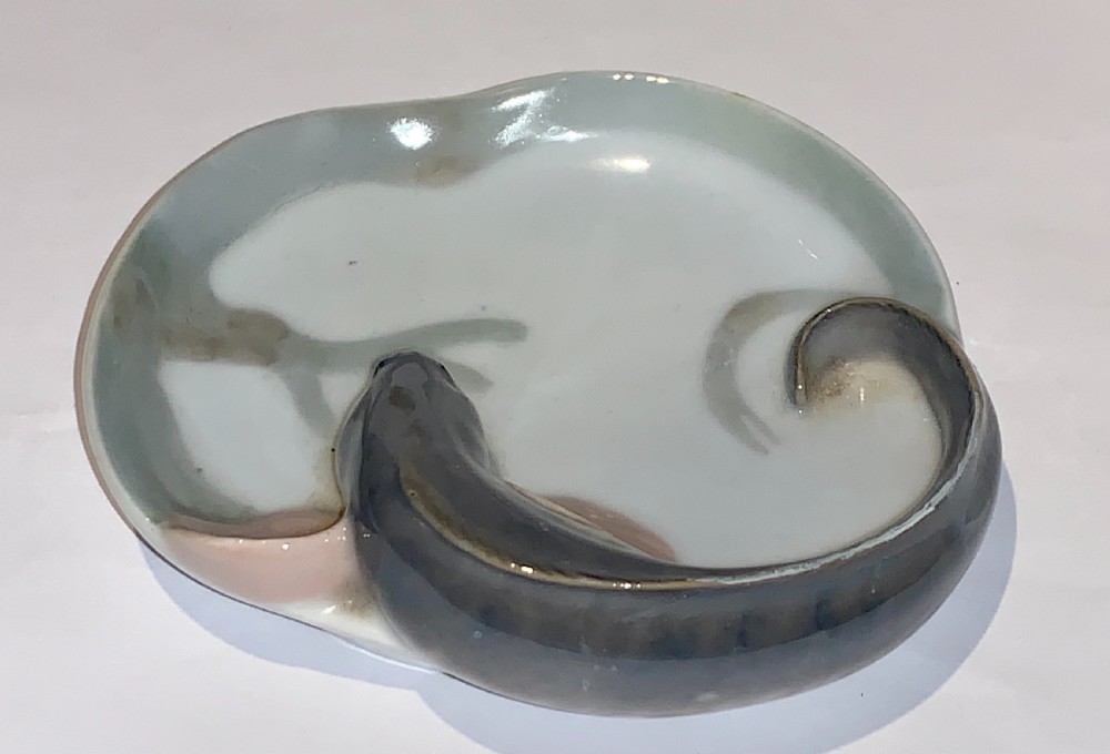 A small circular porcelain dish featuring a sculpted grey fish with pink fins moulded along the curved rim against an off-white background. View IV