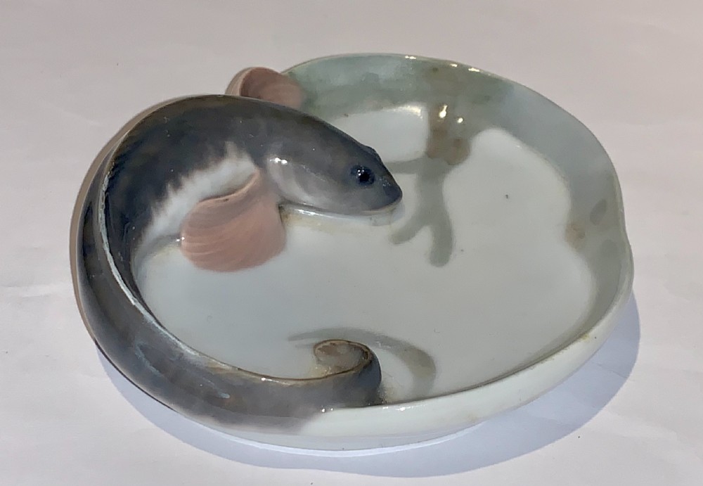 A small circular porcelain dish featuring a sculpted grey fish with pink fins moulded along the curved rim against an off-white background. View V