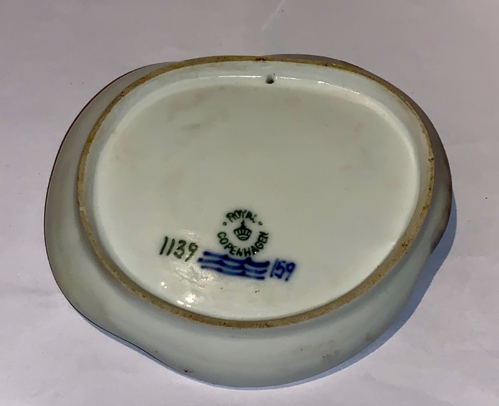 A small circular porcelain dish featuring a sculpted grey fish with pink fins moulded along the curved rim against an off-white background. View VI
