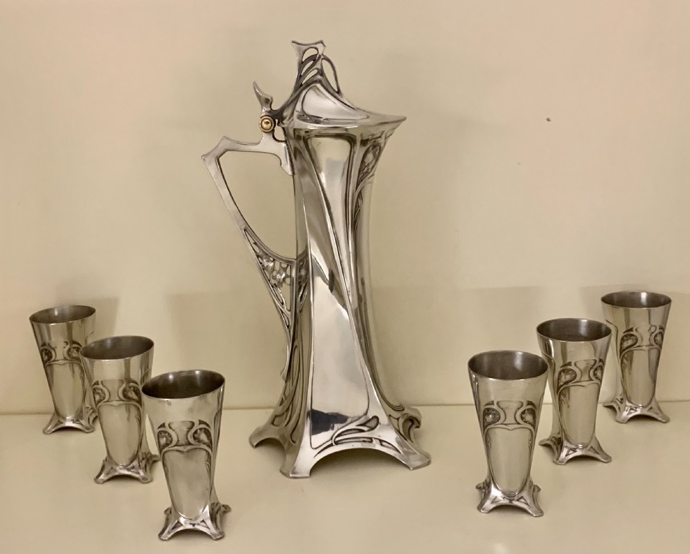 An Art Nouveau silvered pewter liqueur set comprising a tall spouted decanter with organic handles and six matching small tapered cups. View VII