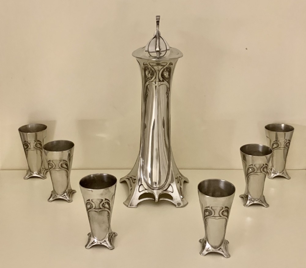 An Art Nouveau silvered pewter liqueur set comprising a tall spouted decanter with organic handles and six matching small tapered cups. View XXIV
