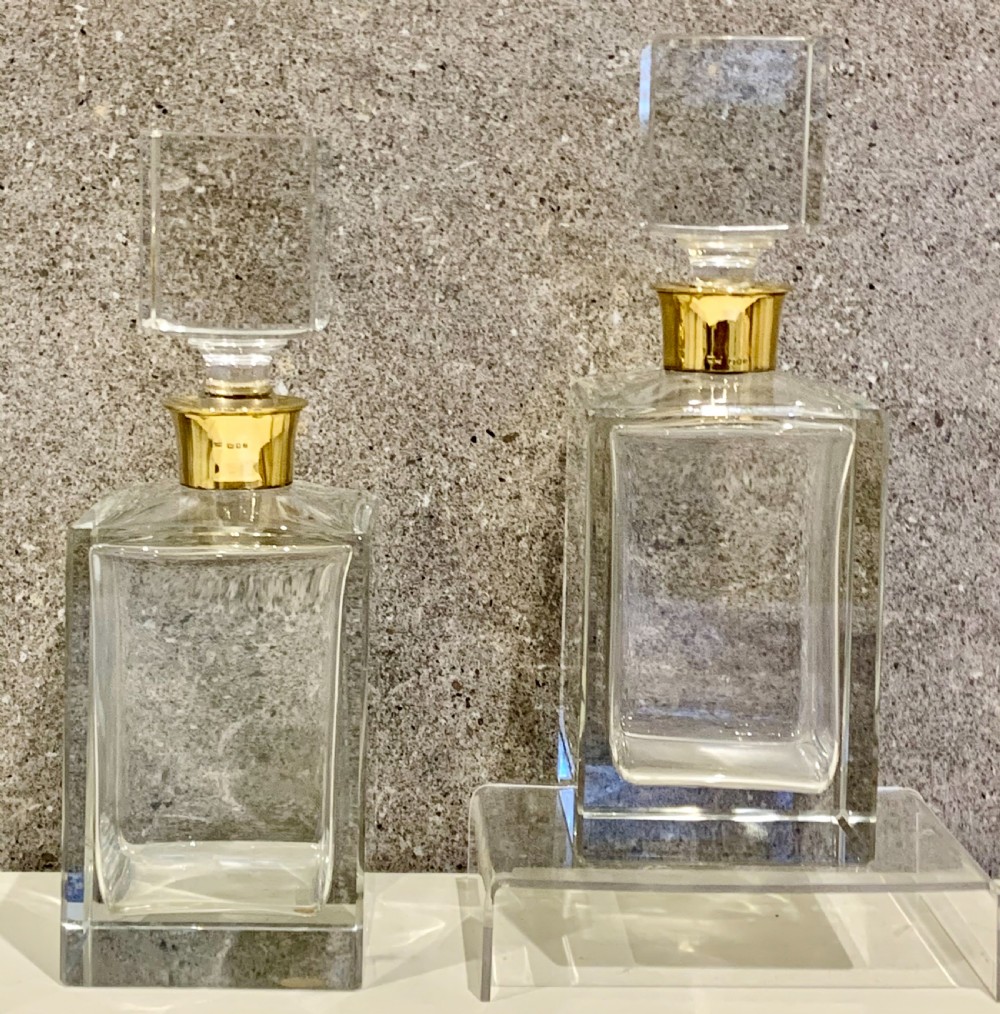 A pair of rectangular clear glass spirit decanters featuring gold-toned silver collars around the necks and matching square glass stoppers. View V