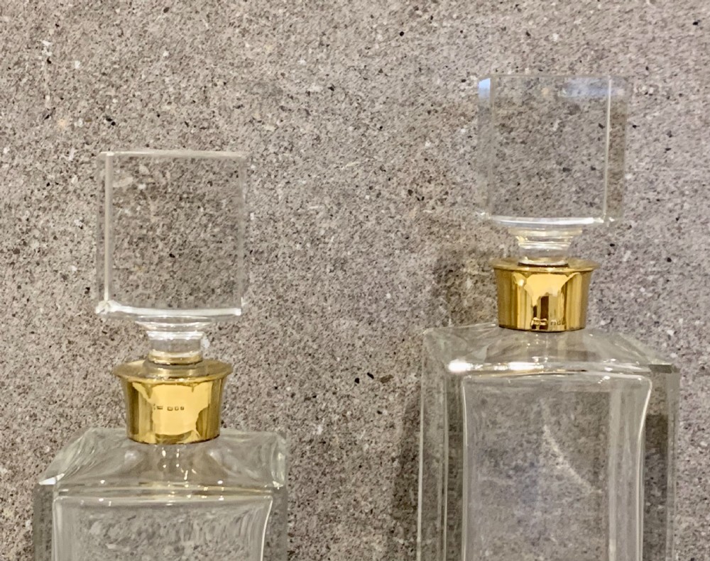 A pair of rectangular clear glass spirit decanters featuring gold-toned silver collars around the necks and matching square glass stoppers. View IX