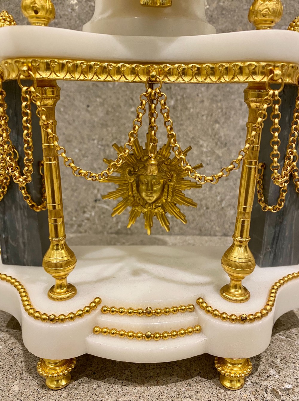 A three-piece Louis XVI style clock garniture set featuring a central white and grey marble clock adorned with gilt bronze mounts and a sunburst pendulum, flanked by two matching marble obelisk candle holders. View VIII