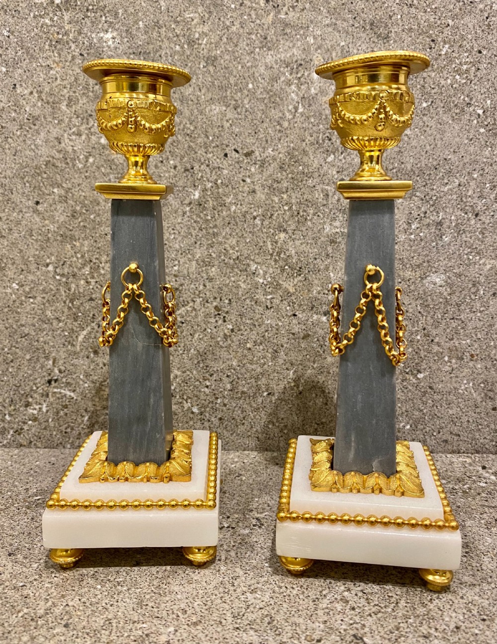 A three-piece Louis XVI style clock garniture set featuring a central white and grey marble clock adorned with gilt bronze mounts and a sunburst pendulum, flanked by two matching marble obelisk candle holders. View XII