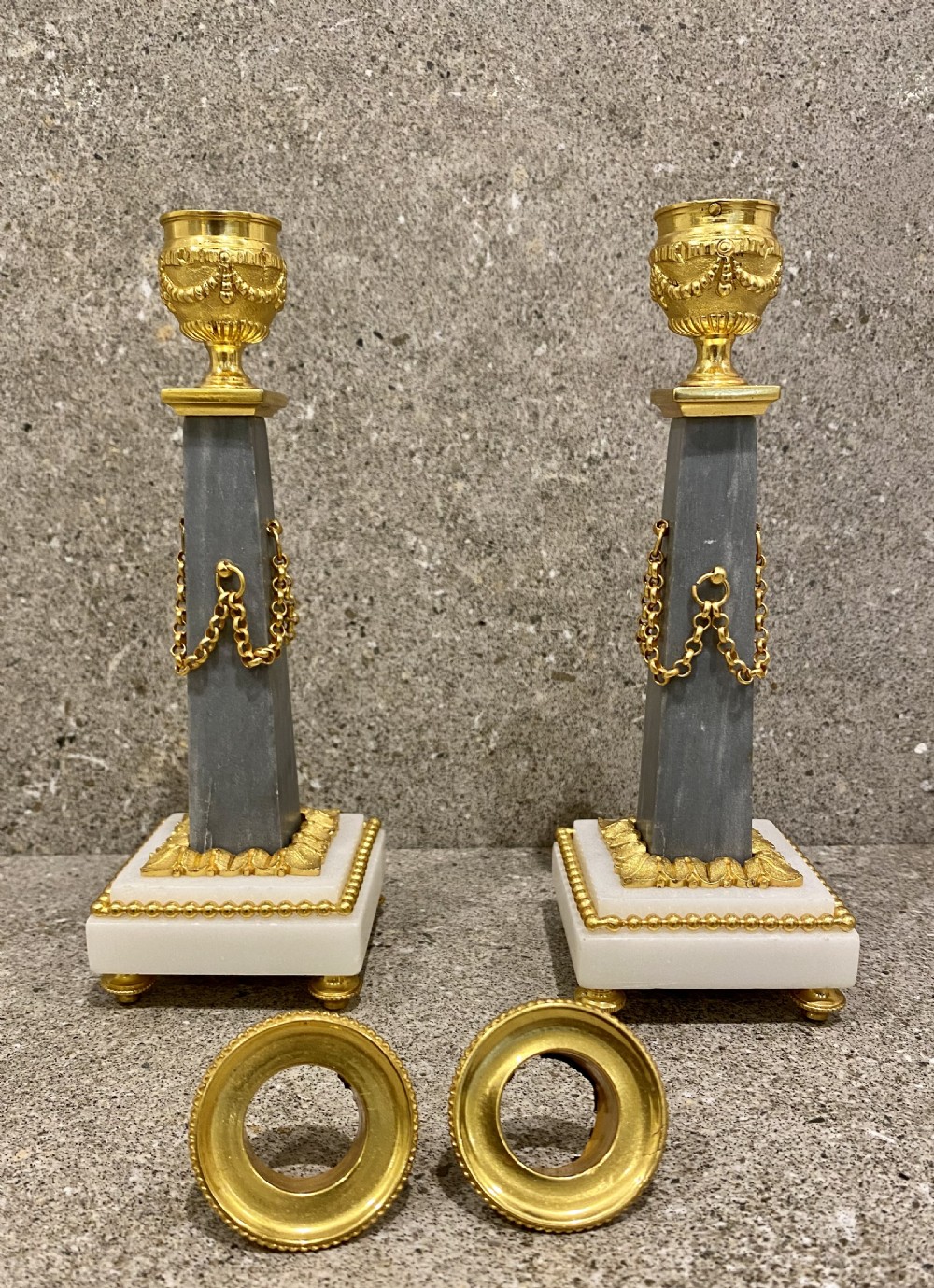A three-piece Louis XVI style clock garniture set featuring a central white and grey marble clock adorned with gilt bronze mounts and a sunburst pendulum, flanked by two matching marble obelisk candle holders. View XIII