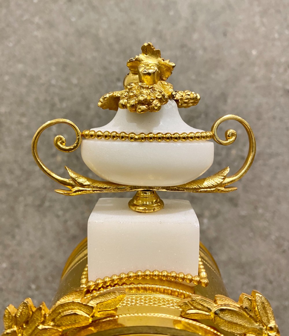 A three-piece Louis XVI style clock garniture set featuring a central white and grey marble clock adorned with gilt bronze mounts and a sunburst pendulum, flanked by two matching marble obelisk candle holders. View XIV