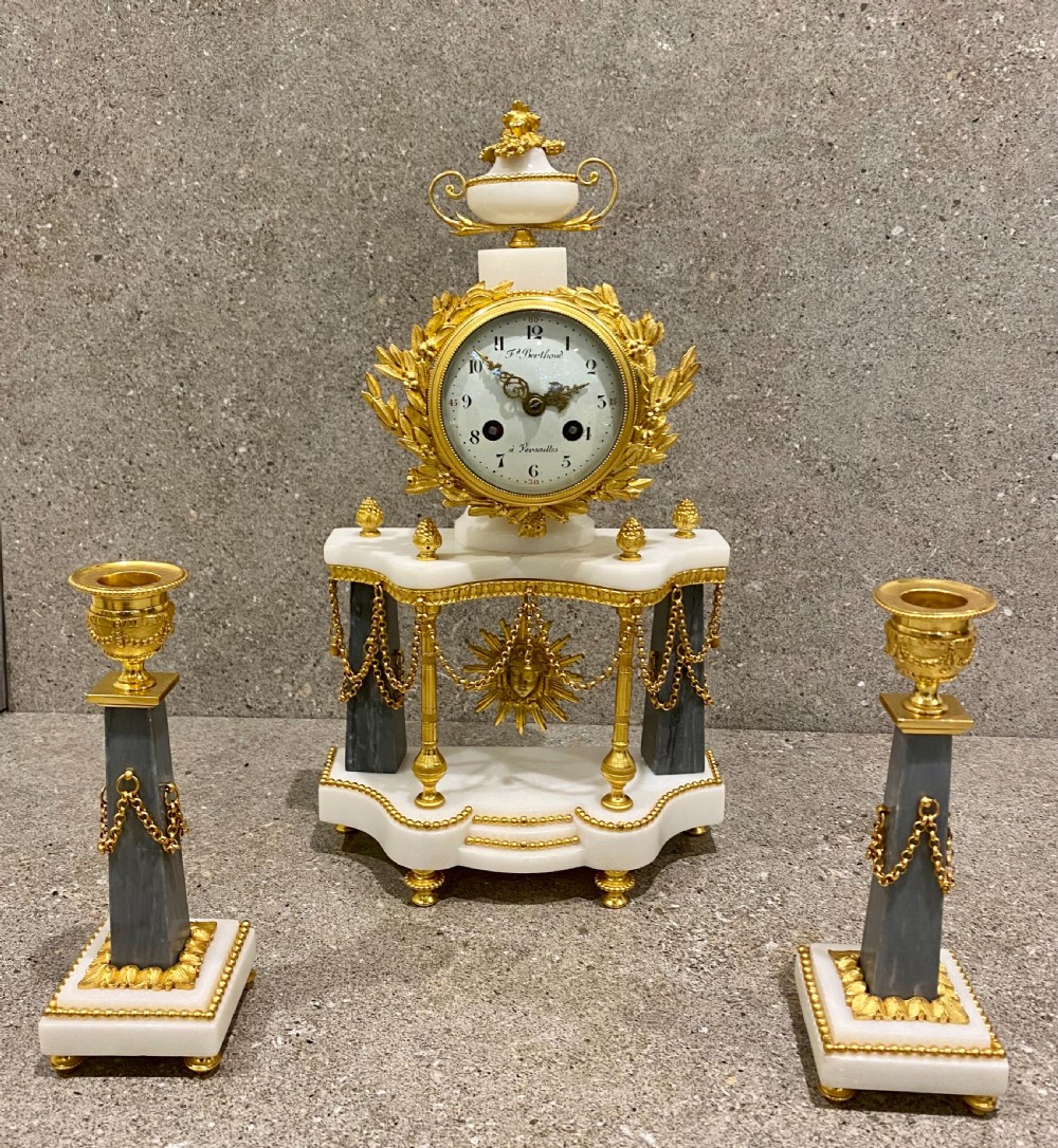 A three-piece Louis XVI style clock garniture set featuring a central white and grey marble clock adorned with gilt bronze mounts and a sunburst pendulum, flanked by two matching marble obelisk candle holders. View XVII