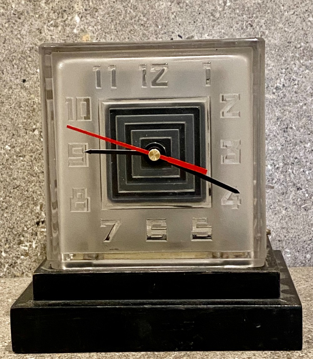 An Art Deco square glass clock featuring raised geometric numerals, a central stepped square motif, and a multi-tiered black base. View II