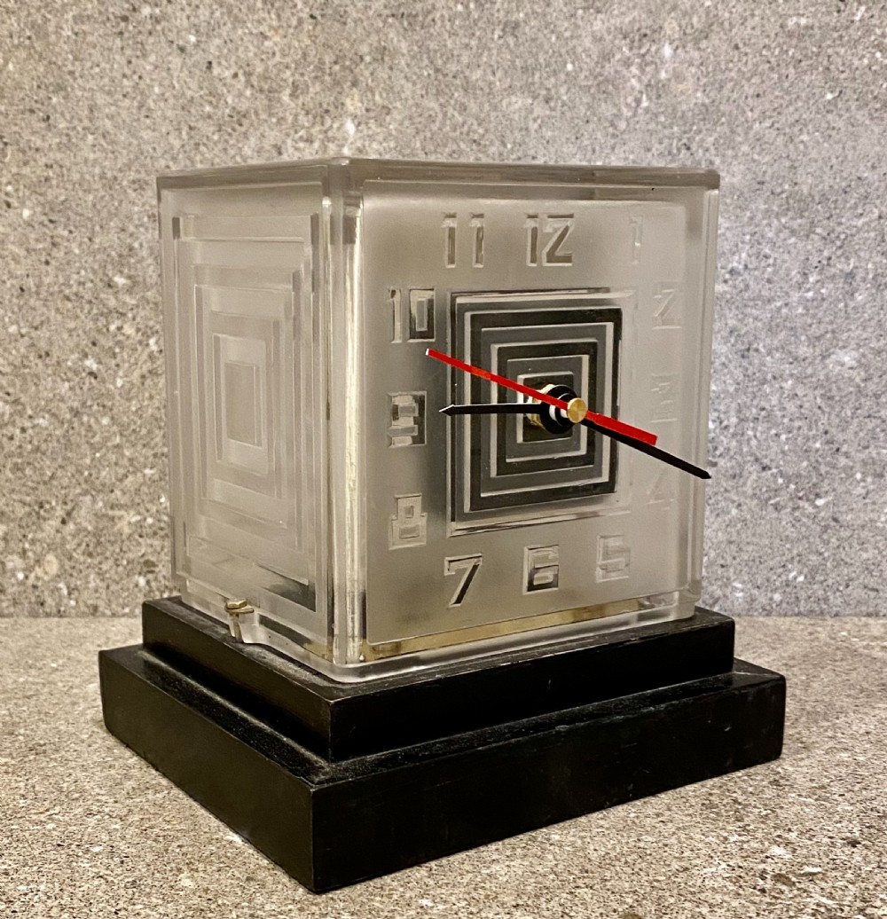 An Art Deco square glass clock featuring raised geometric numerals, a central stepped square motif, and a multi-tiered black base. View III