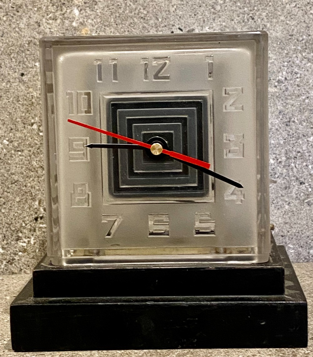 An Art Deco square glass clock featuring raised geometric numerals, a central stepped square motif, and a multi-tiered black base. View VII
