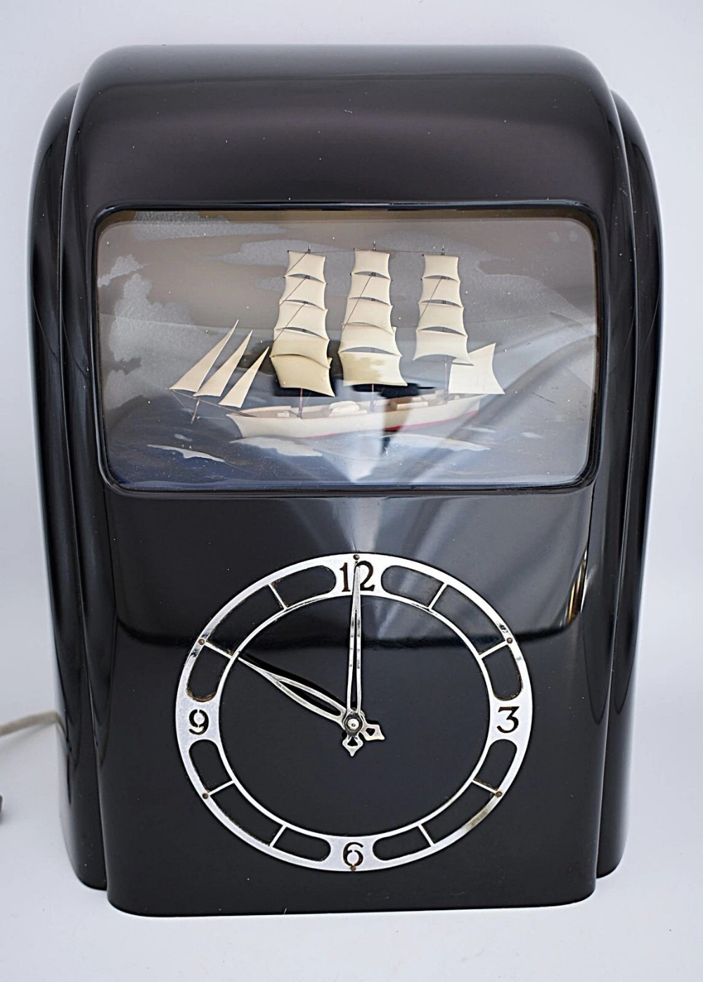 An Art Deco black Bakelite electric clock featuring a chrome chapter ring beneath an illuminated diorama window with a moving ship model.