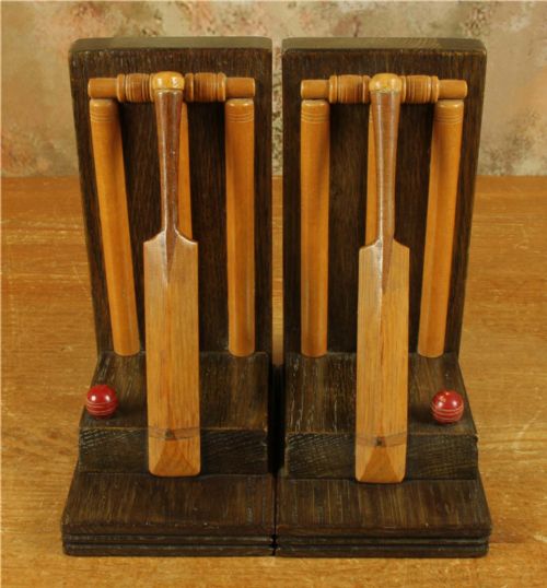 fig 2. A pair of carved dark oak bookends featuring miniature wooden cricket bats, wickets, and red painted cricket balls mounted on stepped rectangular bases.