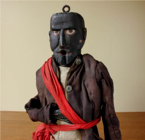 fig 1. An antique carved wooden marionette puppet figure dressed in a dark coat, tan trousers, and a red sash across the body.
