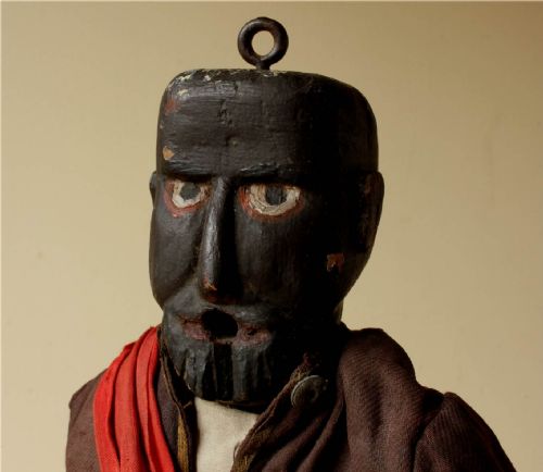 fig 2. An antique carved wooden marionette puppet figure dressed in a dark coat, tan trousers, and a red sash across the body.
