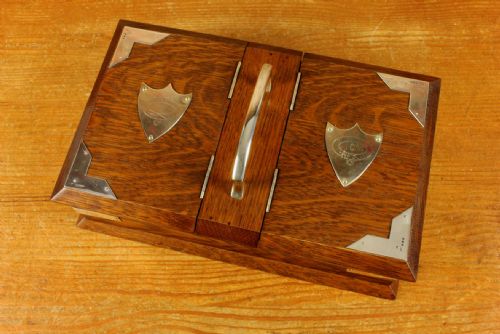 fig 1. A rectangular dark oak wooden box featuring dual hinged lids, a central metal handle, silver corner mounts, and silver shield motifs on each lid.