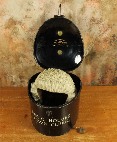 fig 1. An antique legal set comprising a woven horsehair barrister wig on a metal stand beside a cylindrical black tin carrying case with cream lettering reading Eric C. Holmes Town Clerk.
