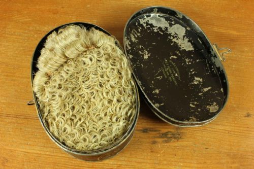 fig 1. A Victorian legal set containing a curled horsehair barrister wig, white cotton court bands, and an oval black metal tin decorated with gilded lettering.