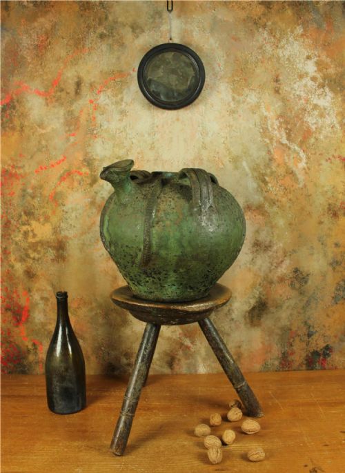 fig 1. A bulbous antique green earthenware jug featuring three handles, a pour spout, and a heavily textured weathered glazed surface.