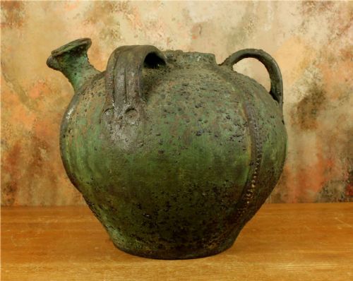fig 2. A bulbous antique green earthenware jug featuring three handles, a pour spout, and a heavily textured weathered glazed surface.