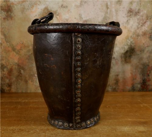fig 1. A dark brown Georgian leather fire bucket featuring a heavy strap handle, brass studs around the base, and a painted royal coat of arms with a lion and unicorn.