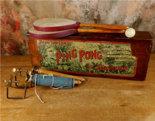 fig 1. An open wooden pine box containing two vellum ping pong rackets, a white ball, and brass net clamps with a folded green net, featuring a Hamleys paper label inside the lid.