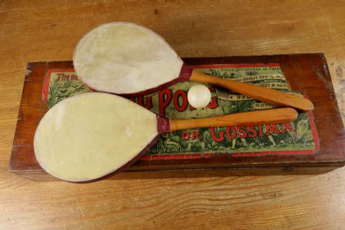 fig 2. An open wooden pine box containing two vellum ping pong rackets, a white ball, and brass net clamps with a folded green net, featuring a Hamleys paper label inside the lid.