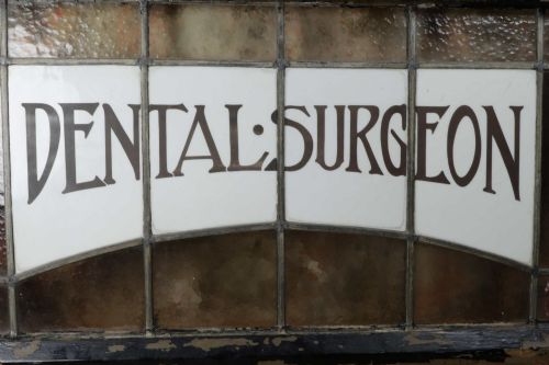 fig 1. An Edwardian leaded glass window panel set in a dark wooden frame with white glass panes featuring black lettering that reads DENTAL SURGEON.