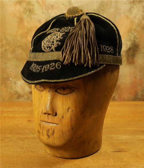 fig 1. A 1920s black velvet sports cap featuring metallic braid piping, embroidered dates, monogram crest, and a prominent hanging tassel.