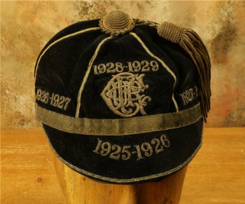 fig 2. A 1920s black velvet sports cap featuring metallic braid piping, embroidered dates, monogram crest, and a prominent hanging tassel.
