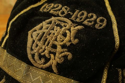 fig 3. A 1920s black velvet sports cap featuring metallic braid piping, embroidered dates, monogram crest, and a prominent hanging tassel.