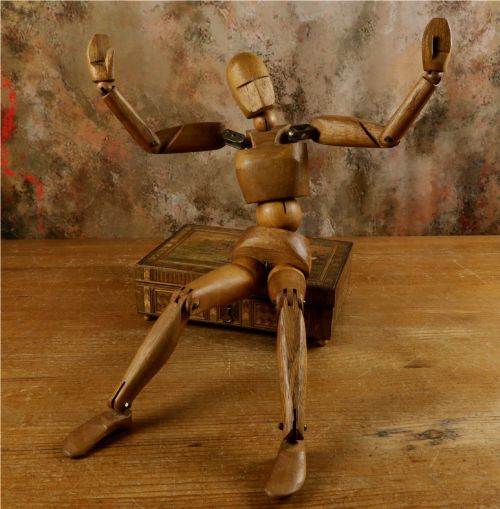 fig 1. A carved teak articulated artist lay figure standing on a surface with both arms raised at the elbow joints.