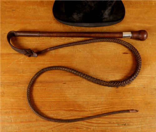 fig 1. A dark brown braided leather hunting whip with a turned knob handle and an engraved silver collar resting coiled on a wooden surface.