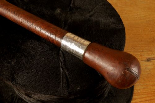 fig 2. A dark brown braided leather hunting whip with a turned knob handle and an engraved silver collar resting coiled on a wooden surface.