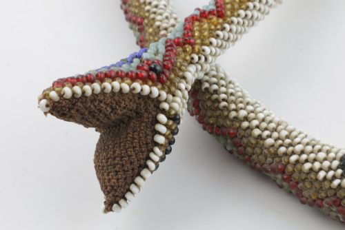 fig 1. A long, hand-crafted glass beadwork snake with white, olive green, and red geometric patterns coiled into a spiral, featuring text reading TURKISH PRISONERS 1918 woven into its body.