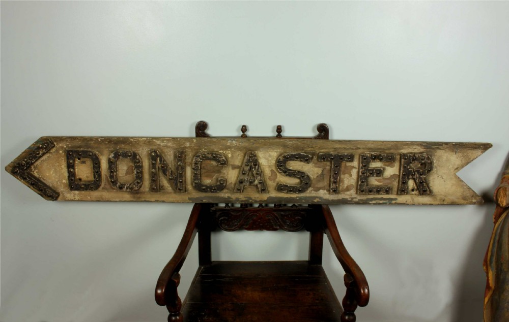 A large, arrow-shaped wooden direction sign painted in weathered cream with the word DONCASTER outlined in raised black lettering set with glass reflectors. View II