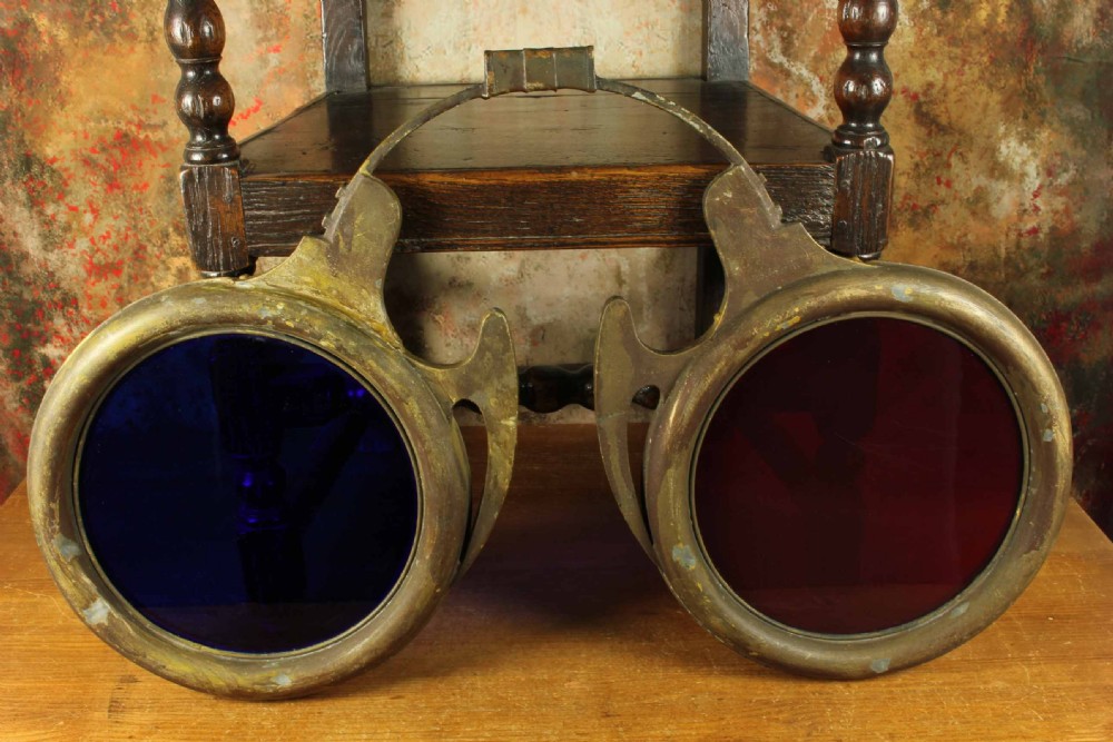 An antique brass optician trade sign in the form of oversized pince-nez spectacles, featuring one red glass lens and one dark blue glass lens. View X