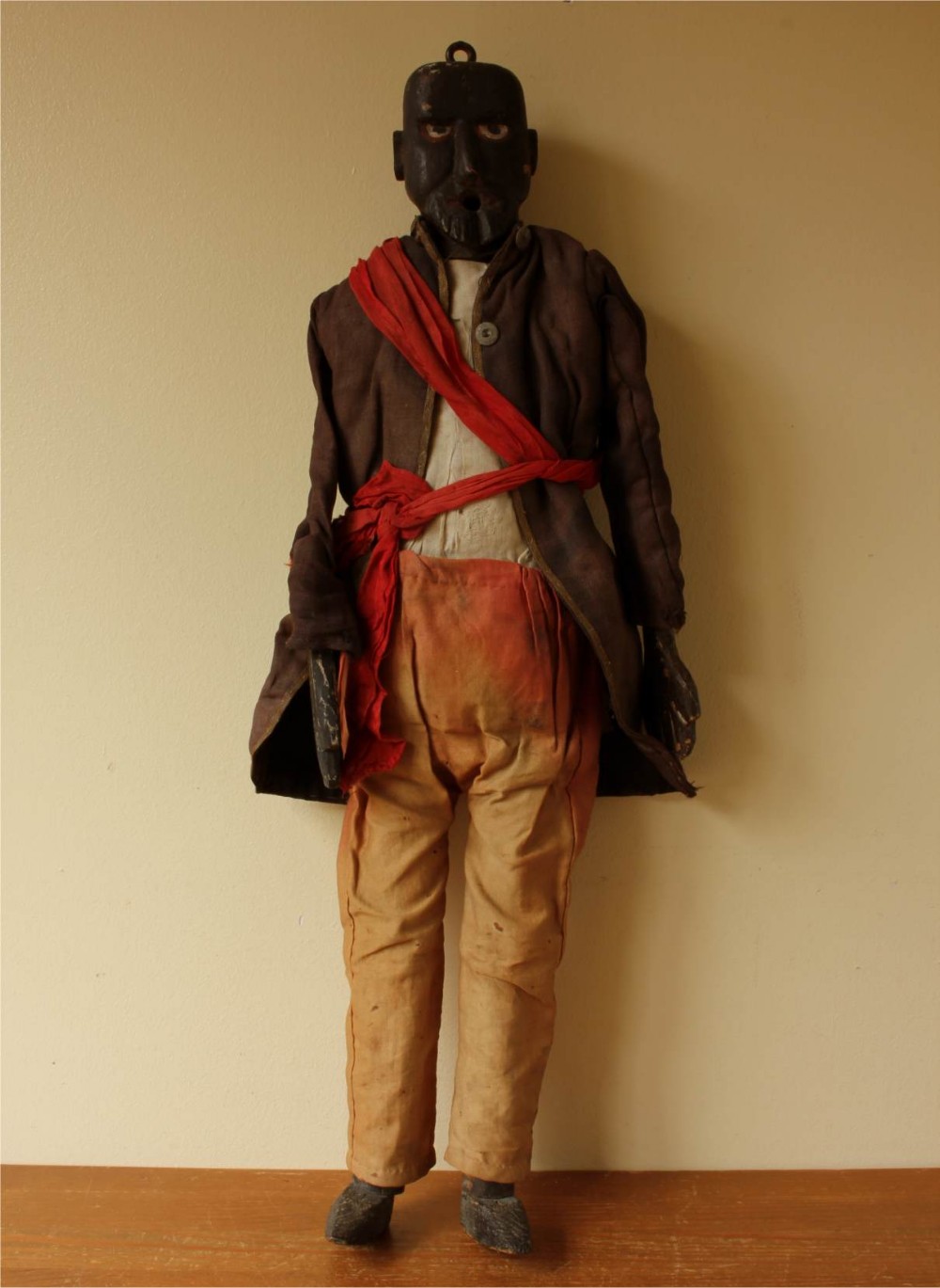 An antique carved wooden marionette puppet figure dressed in a dark coat, tan trousers, and a red sash across the body.