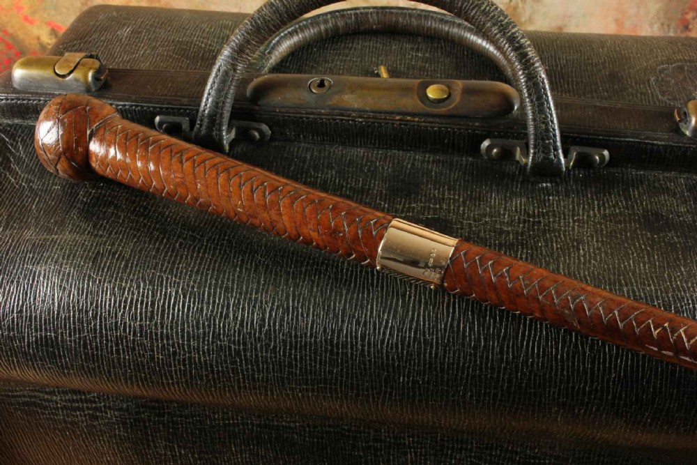 An antique woven dark brown leather riding crop featuring a polished gold collar band and a leather keeper loop at the tip. View V