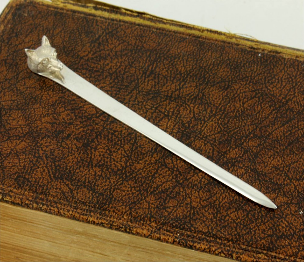 A silver letter opener featuring a sculpted fox head finial atop a smooth, flat tapered blade resting on a brown leather surface.