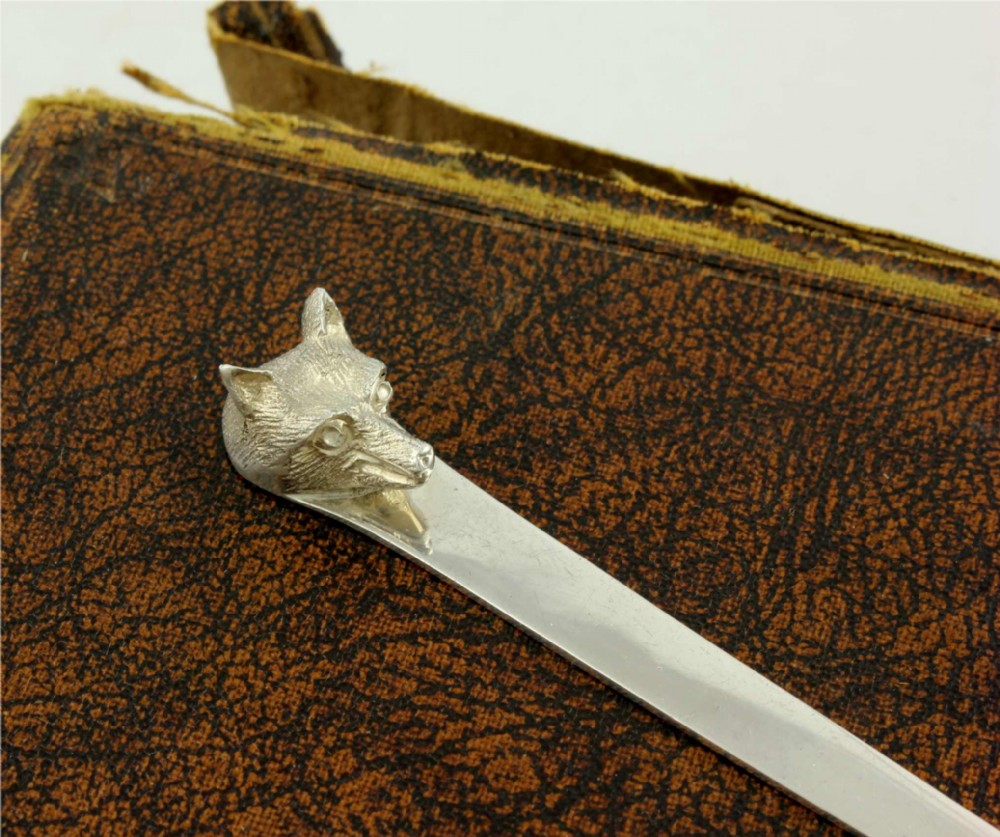 A silver letter opener featuring a sculpted fox head finial atop a smooth, flat tapered blade resting on a brown leather surface. View III