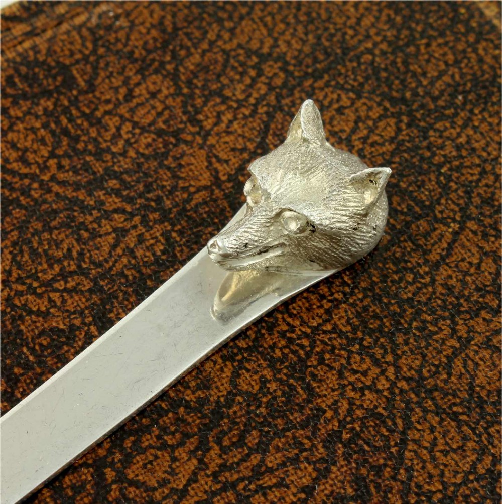A silver letter opener featuring a sculpted fox head finial atop a smooth, flat tapered blade resting on a brown leather surface. View V