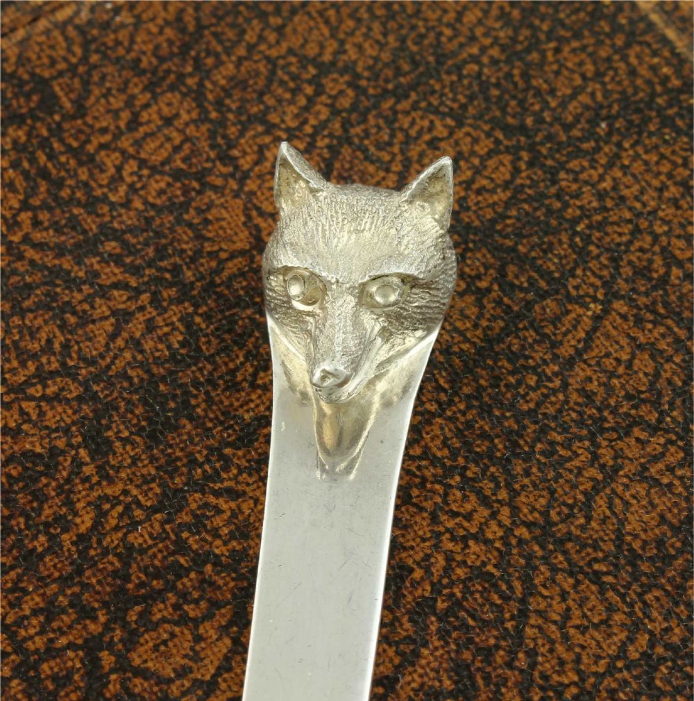 A silver letter opener featuring a sculpted fox head finial atop a smooth, flat tapered blade resting on a brown leather surface. View XI