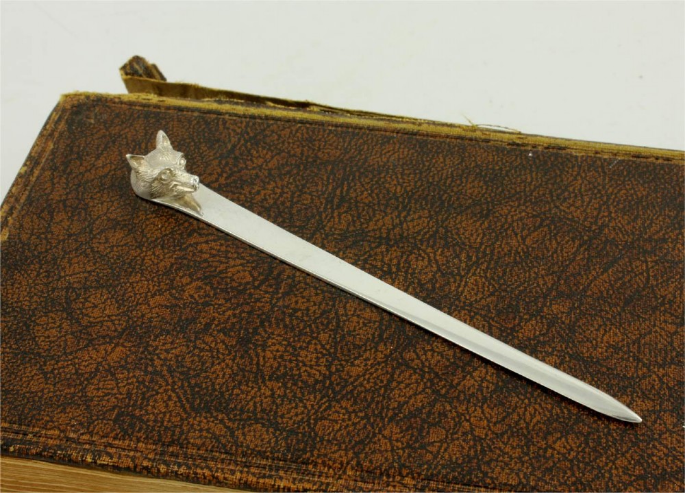 A silver letter opener featuring a sculpted fox head finial atop a smooth, flat tapered blade resting on a brown leather surface. View XII