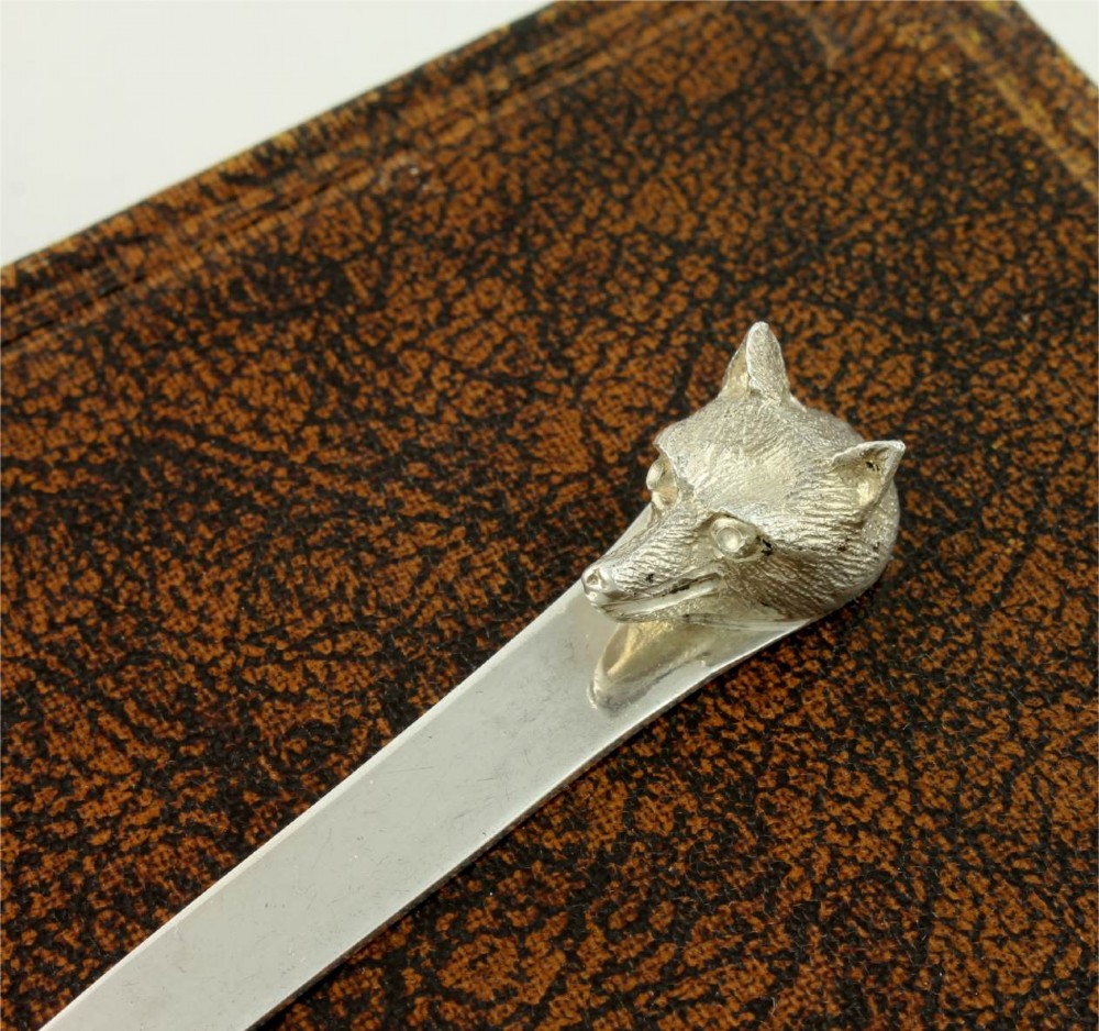 A silver letter opener featuring a sculpted fox head finial atop a smooth, flat tapered blade resting on a brown leather surface. View XIV