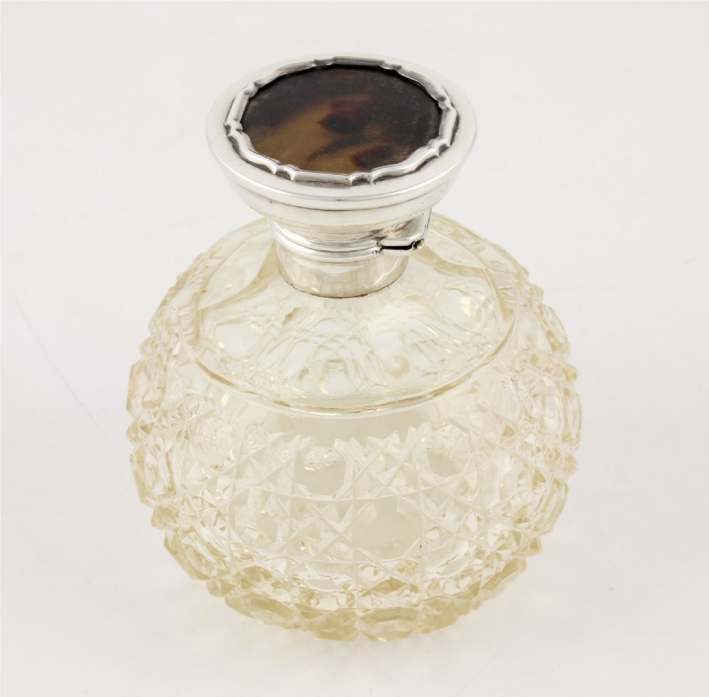 A spherical cut glass scent bottle featuring a hallmarked sterling silver hinged cap inlaid with a round piece of tortoiseshell.