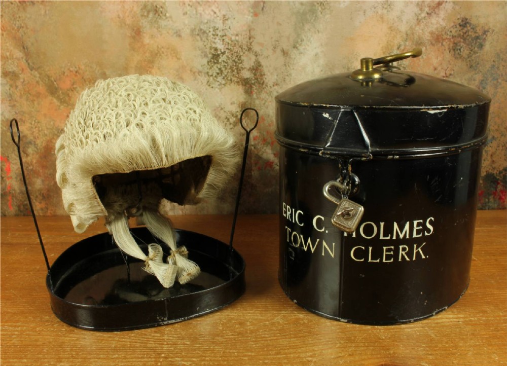 An antique legal set comprising a woven horsehair barrister wig on a metal stand beside a cylindrical black tin carrying case with cream lettering reading Eric C. Holmes Town Clerk.