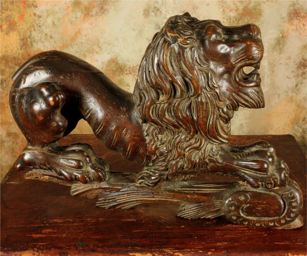 A carved dark wood lion sculpture shown in a reclining position with an open mouth and detailed mane.